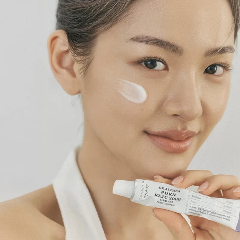 Woman applying Dr.Althea PDRN Reju 2000 Cream on her cheek, sold at bareskin.lt, showing the product tube packaging.