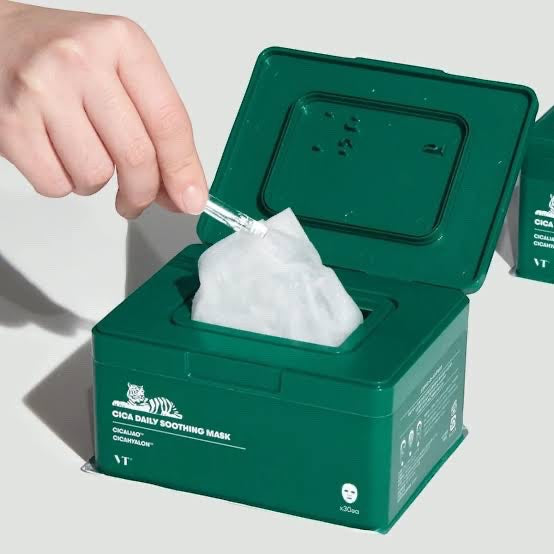 Hand pulling a thin sheet mask from a green VT Cosmetics Cica Daily Soothing Mask dispenser box for skincare.